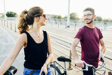 Couple talking while walking with bicycles on bridge against clear sky - SBAF00025