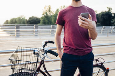 Nahaufnahme eines Mannes, der ein Smartphone benutzt, während er mit dem Fahrrad auf einer Brücke bei Sonnenuntergang steht - SBAF00026