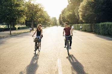 Glückliches Paar Radfahren auf der Straße inmitten von Bäumen im Park bei Sonnenuntergang - SBAF00029