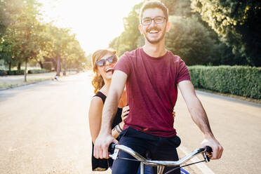 Fröhlicher Mann mit Freundin auf dem Fahrrad auf der Straße im Park bei Sonnenuntergang - SBAF00031