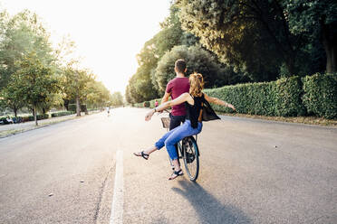 Young woman with arms outstretched sitting with boyfriend on bicycle in park - SBAF00032