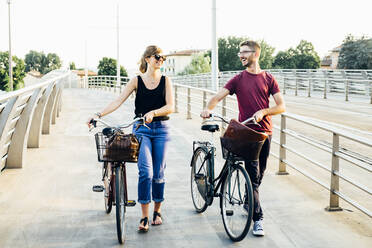 Happy couple talking while walking with bicycles on bridge against clear sky - SBAF00033