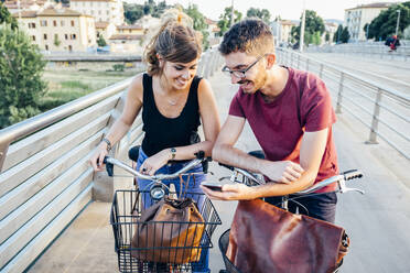 Man showing smart phone to girlfriend while standing with bicycles on bridge - SBAF00036