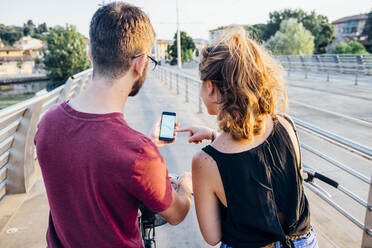 Ein Paar benutzt ein Smartphone, während es mit Fahrrädern auf einer Brücke bei Sonnenuntergang steht - SBAF00037