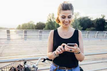 Smiling young woman using smart phone while standing with bicycle on bridge against clear sky - SBAF00038