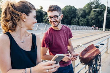 Happy couple using smart phones while standing on bridge at sunset - SBAF00039