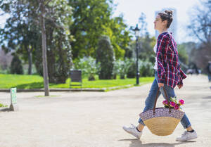 Seitenansicht einer jungen Hipster-Frau, die einen Weidenkorb hält, während sie an einem sonnigen Tag in einem Park spazieren geht - ADSF02896