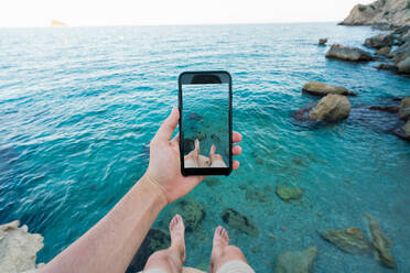 Ausschnittaufnahme eines Mannes, der sein Smartphone benutzt und ein Foto von seinen Beinen macht, während er auf einer Klippe über dem türkisfarbenen Wasser des Meeres sitzt - ADSF02902