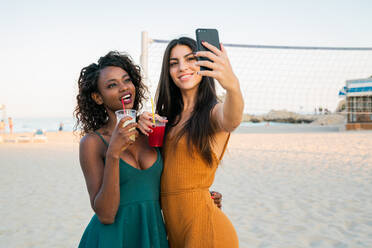 Schöne, trendige, multiethnische Frauen, die Getränke zu sich nehmen und mit dem Handy ein Selfie machen, während sie gemeinsam am Strand chillen - ADSF02912