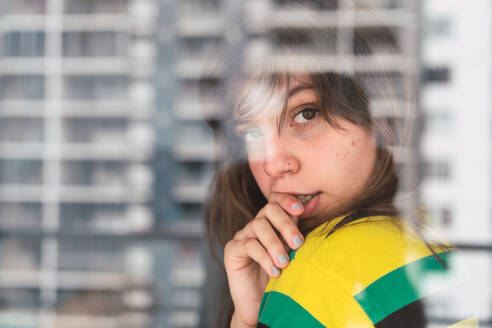 Attraktive junge Frau, die in den Finger beißt und in die Kamera schaut, während sie sich zu Hause an eine Fensterscheibe lehnt - ADSF02917