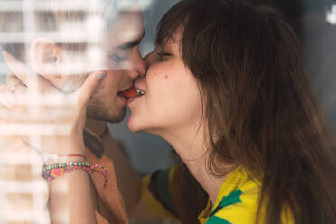 Side view of handsome young man and lovely woman kissing passionately while standing near window at home - ADSF02918