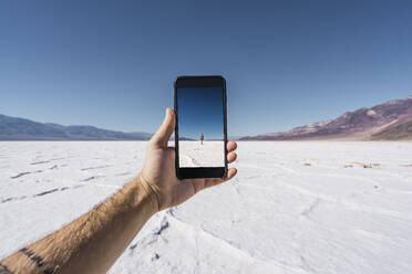 Männlich, der die Hand ausstreckt und das Handy in der Hand hält, mit einem Foto von einer Person in der Nähe eines wilden Feldes mit hellem Boden zwischen Hügeln und einem schönen blauen Himmel - ADSF02930