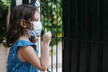 Close-up of girl wearing mask looking through fence while standing in yard - EGAF00476