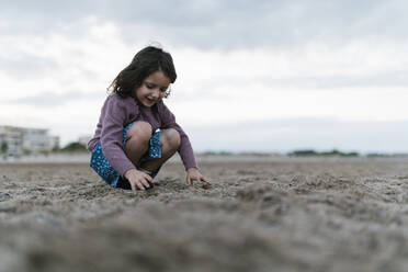 Smiling girl playing with sand while crouching at beach against cloudy sky - EGAF00477