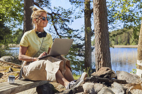 Mittlere erwachsene Frau mit Sonnenbrille, die einen Laptop benutzt, während sie im Tiveden National Park, Schweden, sitzt - UKOF00011