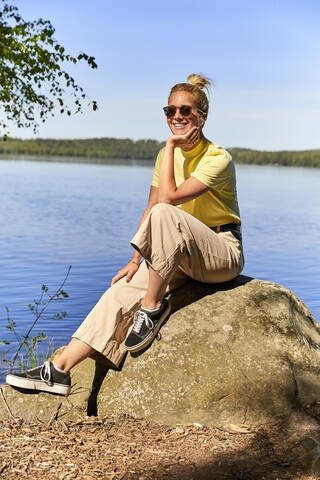 Lächelnde Frau mit Sonnenbrille auf einem Felsen am See im Tiveden National Park, Schweden, lizenzfreies Stockfoto