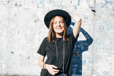 Smiling young woman wearing hat standing against old wall in city on sunny day - MEUF01556