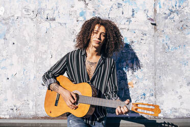 Gut aussehender Mann mit lockigem Haar spielt Gitarre, während er an einer alten Mauer in der Stadt an einem sonnigen Tag steht - MEUF01558