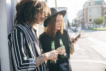 Paar, das Bierflaschen hält und ein Smartphone benutzt, während es auf der Straße in der Stadt steht - MEUF01574