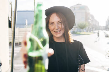 Nahaufnahme einer lächelnden Frau, die eine Bierflasche hält, während sie an einem sonnigen Tag in der Stadt steht - MEUF01575