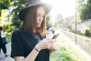 Nahaufnahme einer schönen Frau mit Hut, die im Park ein Smartphone benutzt - MEUF01581