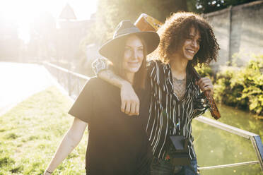 Cheerful man holding guitar with hand on girlfriend's shoulder walking in park during sunny day - MEUF01584