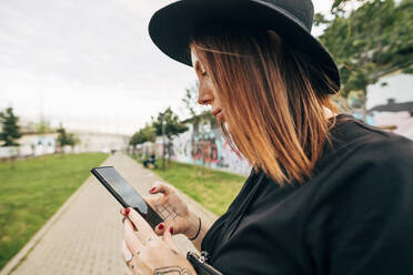 Nahaufnahme einer jungen Frau mit Hut, die im Park ihr Smartphone benutzt - MEUF01602