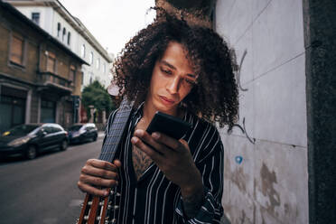 Nahaufnahme eines Mannes mit lockigem Haar, der eine Gitarre hält, während er ein Smartphone in der Stadt benutzt - MEUF01605