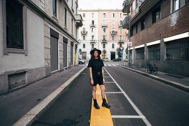Junge Frau mit Hut steht auf der Straße inmitten von Gebäuden in der Stadt - MEUF01606