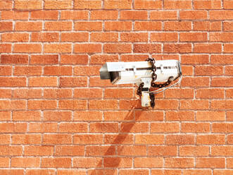 Weiße Überwachungskamera von unten, die mit einer Kette an einer roten Backsteinmauer im Sonnenlicht befestigt ist - ADSF02970