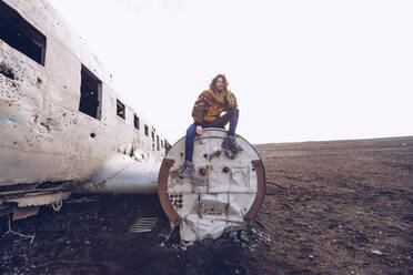 Junge Frau in warmer Kleidung sitzt auf einem kaputten Flugzeug zwischen dunklen Böden in Island - ADSF02974