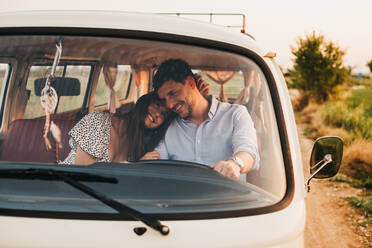 Cheerful young man and woman embracing and riding in vintage van on road in rural environment - ADSF02998