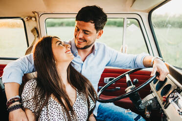 Cheerful young man and woman embracing and riding in vintage van on road in rural environment - ADSF02999