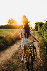 Back view of pretty woman carrying bicycle and walking on rural road in backlit - ADSF03007