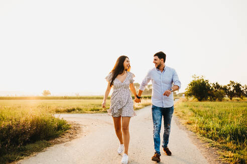 Fröhlicher junger Mann und Frau, die sich an den Händen halten und auf einem Feld im Sonnenlicht laufen - ADSF03008