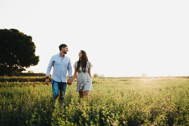 Cheerful young man and woman holding hands and walking on field in sunlight - ADSF03010
