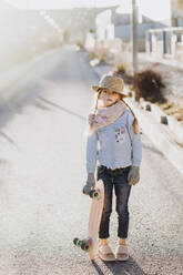 Cute little girl in trendy outfit holding skateboard and looking away while standing on road on sunny day on suburban street - ADSF03068