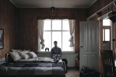 Erwachsener Mann sitzt und ruht sich auf dem Bett aus, während er zu Hause in Island sein Smartphone benutzt - ADSF03142