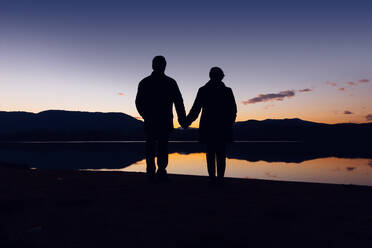 Silhouetten eines nicht erkennbaren Mannes und einer Frau, die sich an den Händen halten, während sie am Ufer in der Nähe des ruhigen Wassers bei herrlichem Sonnenuntergang stehen - ADSF03150