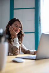 Attractive female charmingly smiling and looking at modern laptop while sitting at office desk near window - ADSF03183