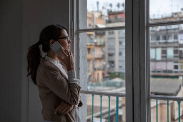 Side view of adult female in glasses talking on phone and looking out window while standing in office - ADSF03185