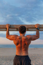 Bärtiger Mann ohne Hemd klettert auf eine horizontale Holzleiter, während er am Strand trainiert - ADSF03207