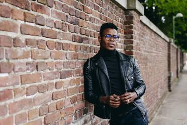 Young handsome african male in glasses and leather jacket standing on brick wall background - ADSF03210