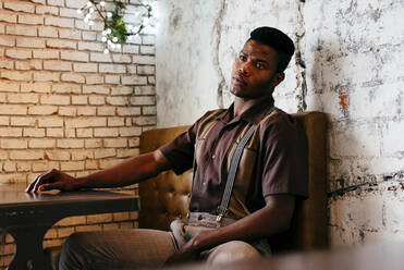 Young black man in pants with suspenders and shirt sitting on couch and looking at camera - ADSF03218