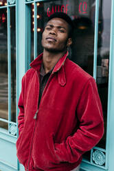 Stilvoller afrikanischer Mann, der seine Hände in die Taschen seines roten Pullovers steckt, steht am Eingang eines Cafés - ADSF03226