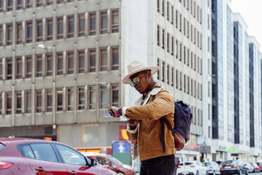 Junger afrikanischer Mann mit Hut und Brille steht an der Straße und schaut in die Wüste - ADSF03227