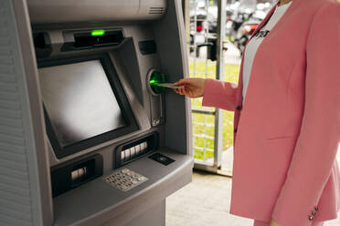 Gesichtslose Aufnahme einer Frau im Anzug, die eine Kreditkarte in einen Geldautomaten steckt, der auf der Straße steht. - ADSF03261