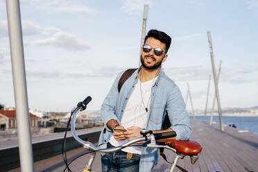 Cheerful hipster man in sunglasses and headphones holding smartphone and standing with retro bicycle on seafront. - ADSF03274