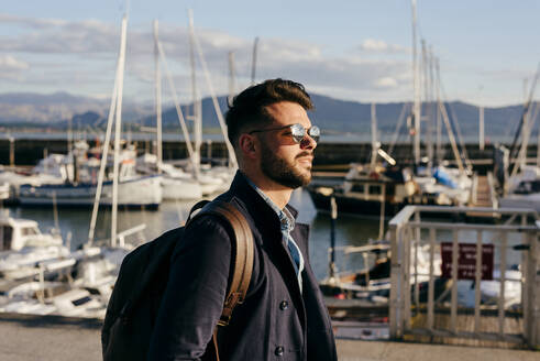 Seitenansicht eines gutaussehenden, trendigen Mannes mit Sonnenbrille und Rucksack vor dem Hintergrund eines Seehafens. - ADSF03279