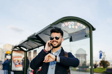 Handsome hipster man in sunglasses checking time and making appointment speaking on phone on bus stop. - ADSF03282